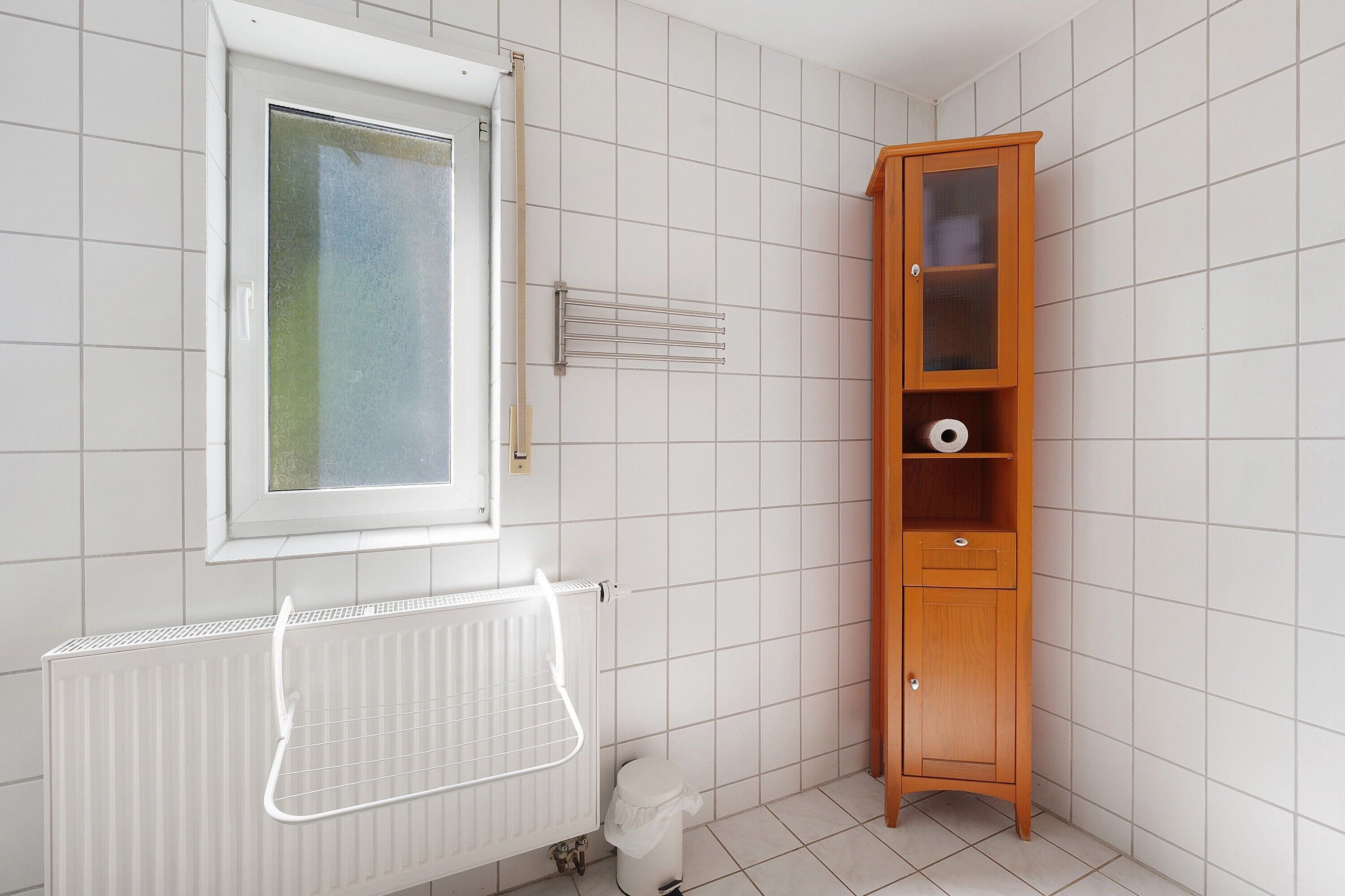 A bathroom with a window, a radiator, a wooden cabinet, and a tiled floor.
