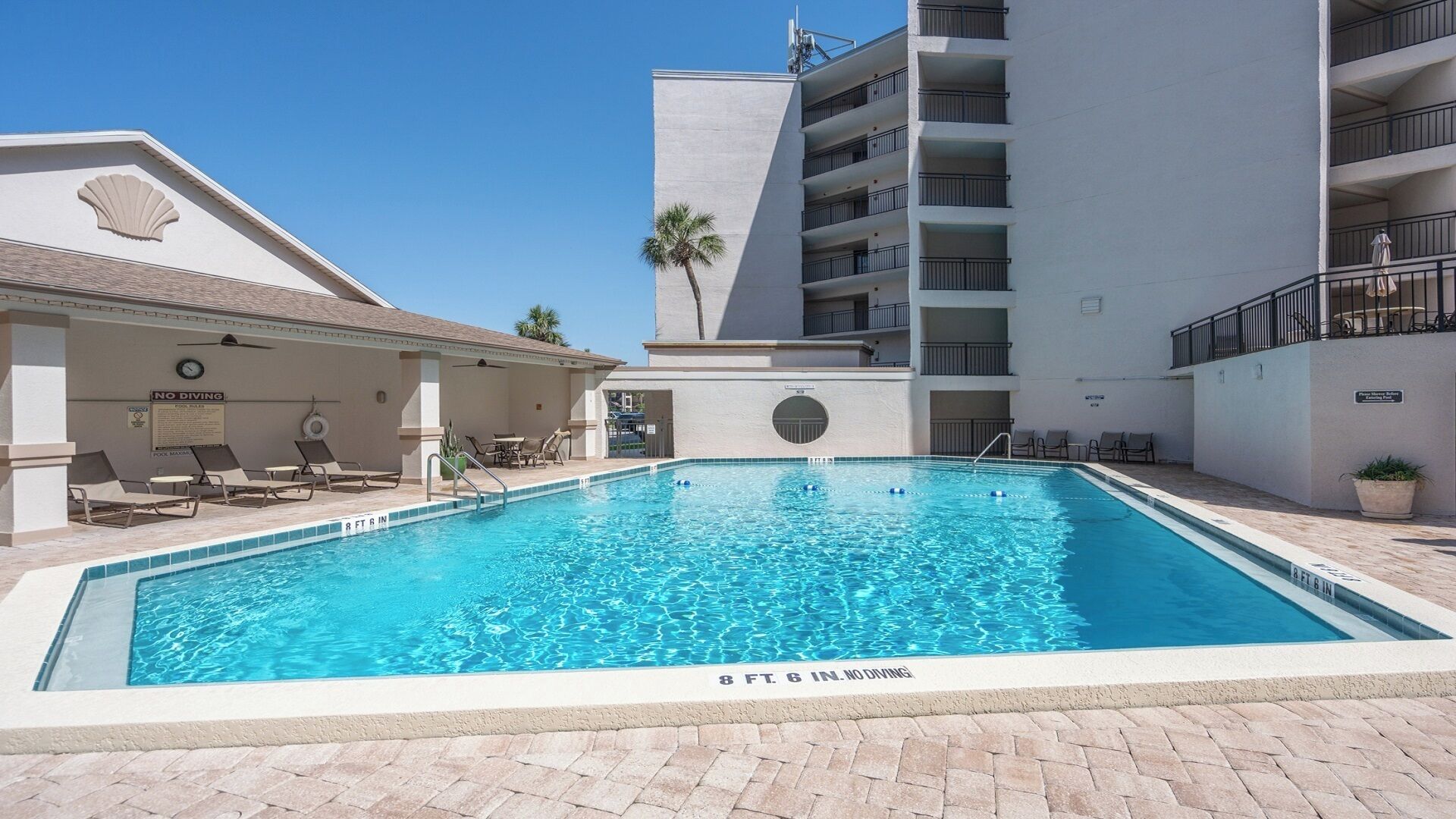 A swimming pool with a sign indicating a depth of 8 feet 6 inches, surrounded by lounge chairs and a building with balconies.