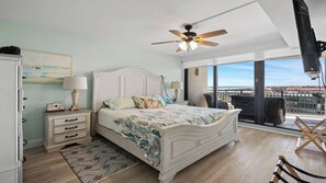 A bedroom with a bed, nightstands, a ceiling fan, a view to the outside, and a balcony with chairs. - Anastasia #605 Ocean front property with panoramic view of ocean/beach, boat parking (St. Augustine Beach)