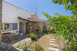 A rustic courtyard with a stone wall, a white building with blue shutters, and a variety of plants. - “Maison De Pays Églantine” with Private Terrace, Garden, and Wi-Fi (Sainte-Marie-de-Ré)