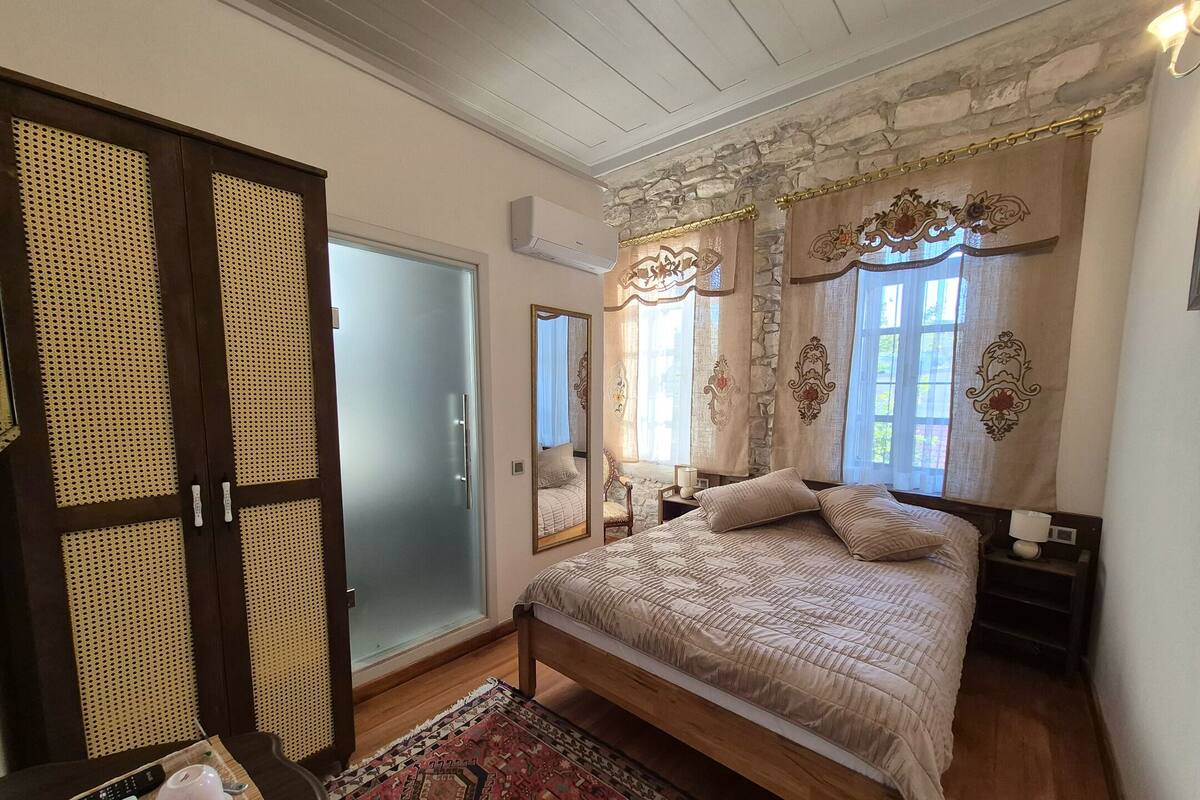 Quarto com cama, guarda-roupa de madeira, espelho, mesa de cabeceira e janela com cortinas.