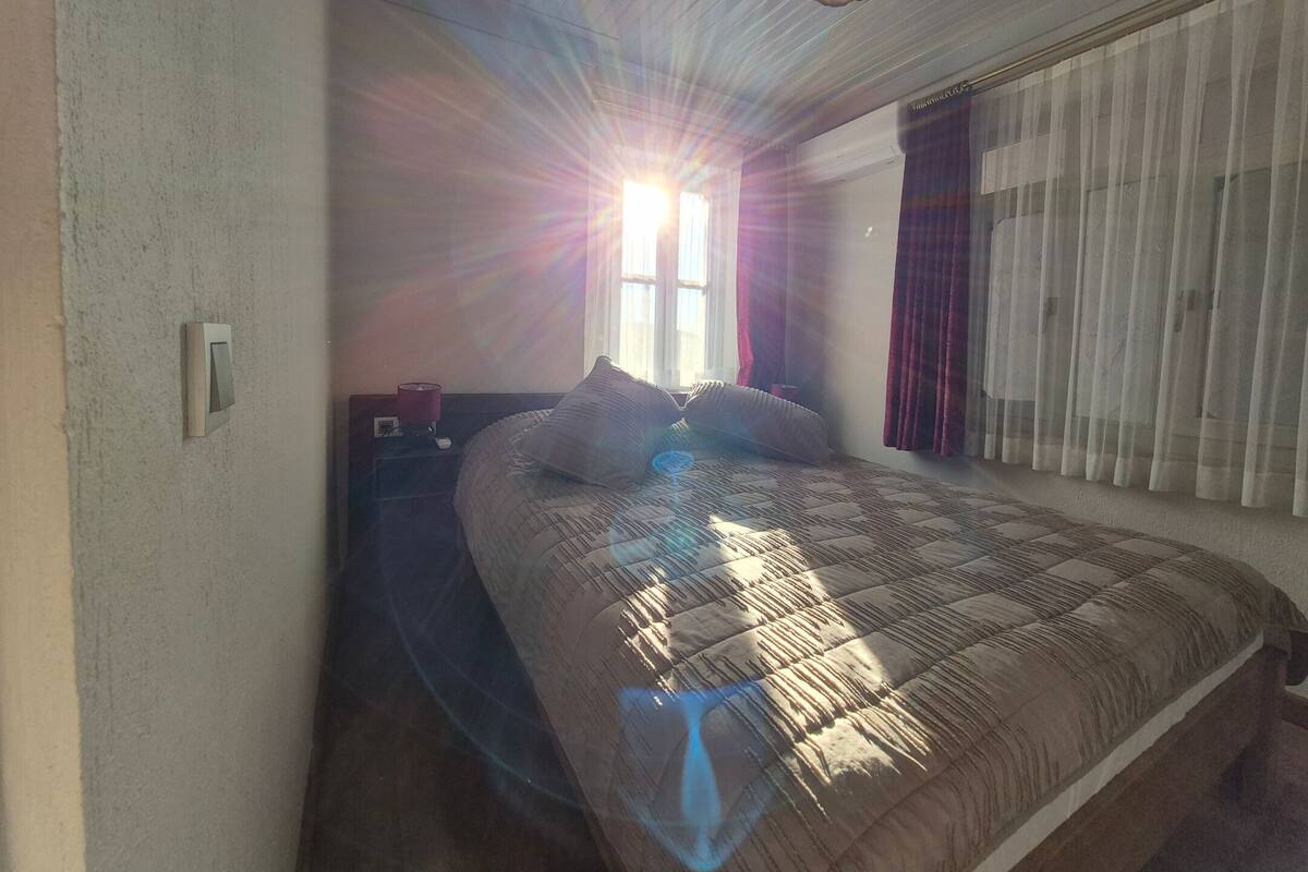 Quarto com cama, mesa de cabeceira, luminária, janela com cortinas e luz no teto.