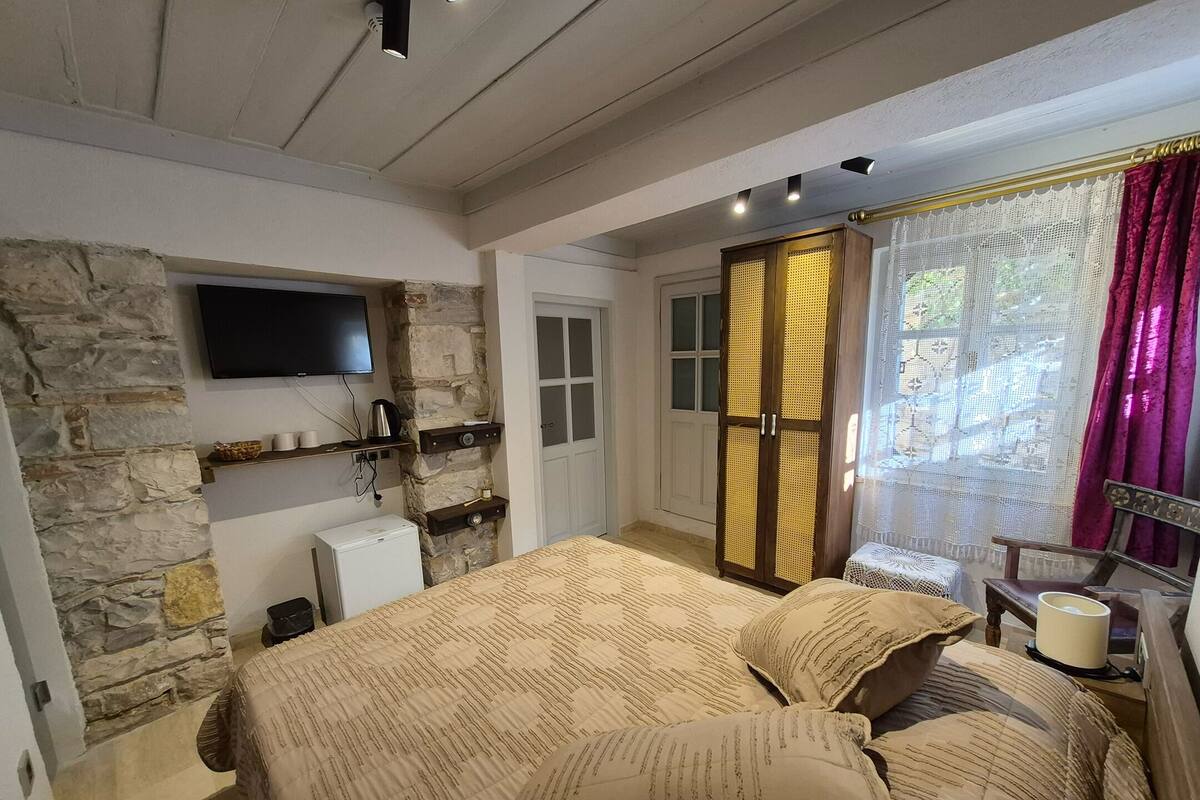 Quarto com parede de pedra, cama com roupa de cama estampada, televisão montada sobre a cama, guarda-roupa com porta trançada e janela com cortinas.
