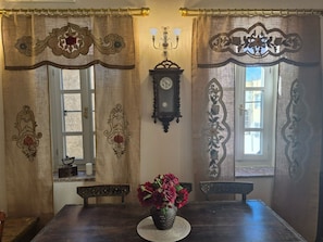 Un comedor con una mesa de madera, dos sillas, un jarrón con flores, un reloj en la pared y cortinas con estampado floral.