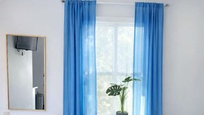Une pièce avec une table ronde, deux chaises, une plante en pot, une fenêtre avec des rideaux bleus et un téléviseur fixé au mur.