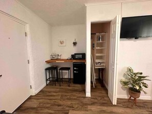 Ein Zimmer mit Küchenzeile, einem Essbereich mit Stühlen, einem an der Wand befestigten Fernseher und einem Blumentopf.