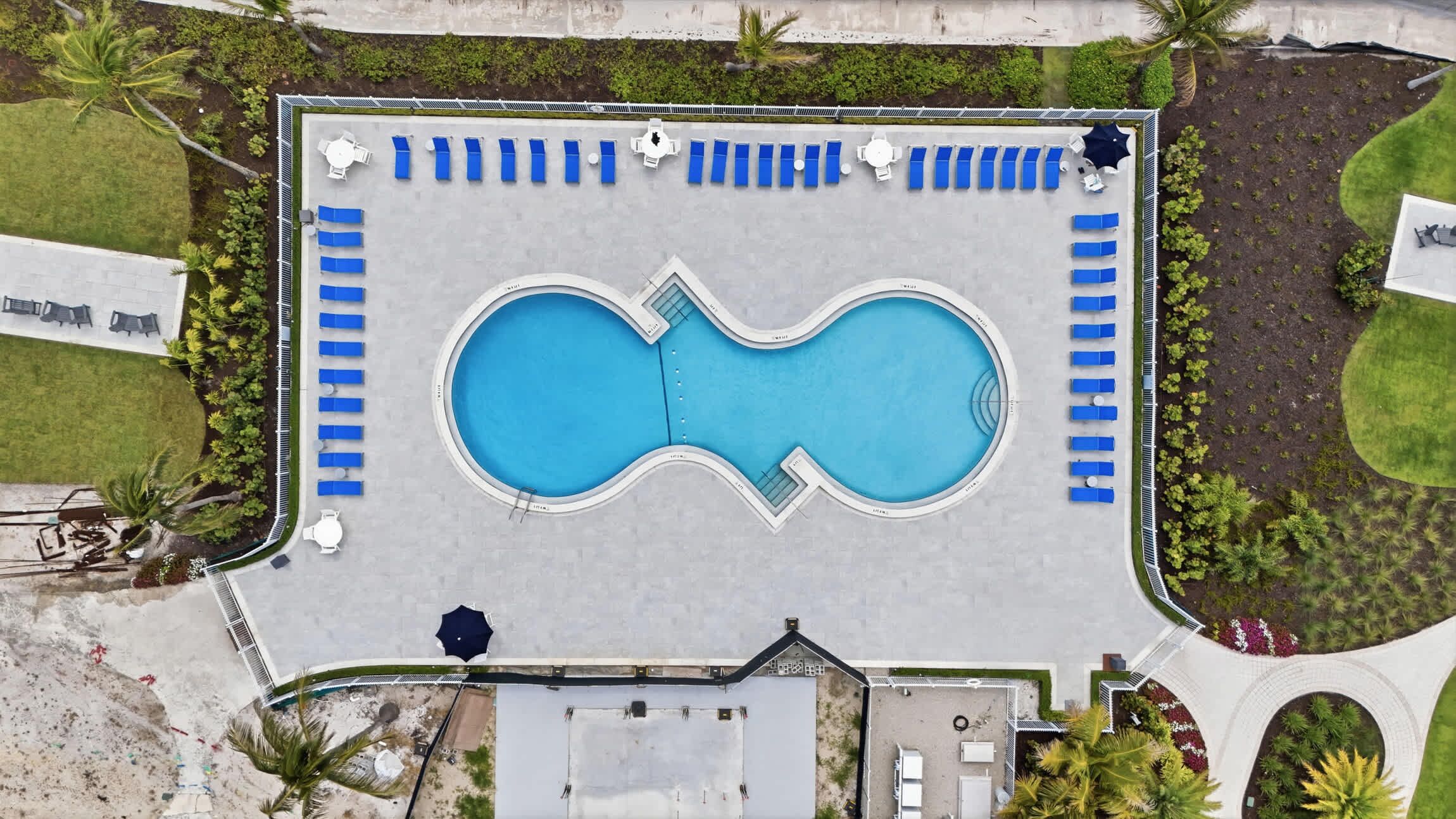 An aerial view of a swimming pool with blue water, surrounded by blue lounge chairs and a few white tables.