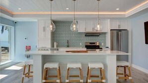 A modern kitchen with white cabinetry, a central island with bar stools, pendant lighting, and a stainless steel refrigerator. - Your Front-Row Seat to Gulf Sunsets on Bonita Beach (Bonita Springs)