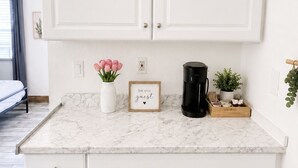 Una cocina con armarios blancos, encimera de mármol, cafetera, planta pequeña y un cartel enmarcado.