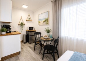 Eine kleine Küchenzeile mit Mikrowelle, Kaffeemaschine und einem kleinen Mülleimer. Ein Esstisch mit zwei Stühlen, ein kleiner Topfpflanze und eine Kerze stehen dort. An der Wand hängt ein Bild mit Strandmotiv, und ein Fenster mit Vorhängen ist zu sehen.