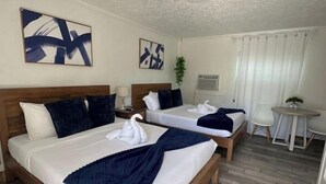 Quarto de hotel com duas camas, cabeceiras de madeira, roupa de cama branca, arte abstrata na parede, uma mesinha e um vaso com planta.