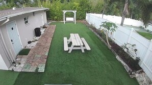 A backyard with artificial grass, a wooden picnic table, a white fence, and a house with a door and windows. - Studio 3 miles from the Beach, with pool #512 (Belleair)