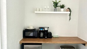 Una kitchenette equipada con microondas, cafetera y un refrigerador pequeño. Encima de ella, un estante blanco con tazas y una planta.