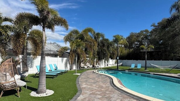 Una piscina de borde curvo, rodeada por un camino pavimentado, palmeras y sillones de descanso.