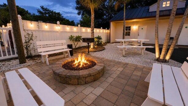 Un patio al aire libre con un hogar para fuego, bancos y mesas blancos.