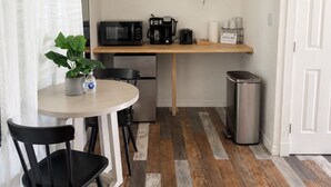 Una kitchenette compacta con microondas, cafetera y un refrigerador pequeño. Hay una mesa con sillas y una planta en maceta.