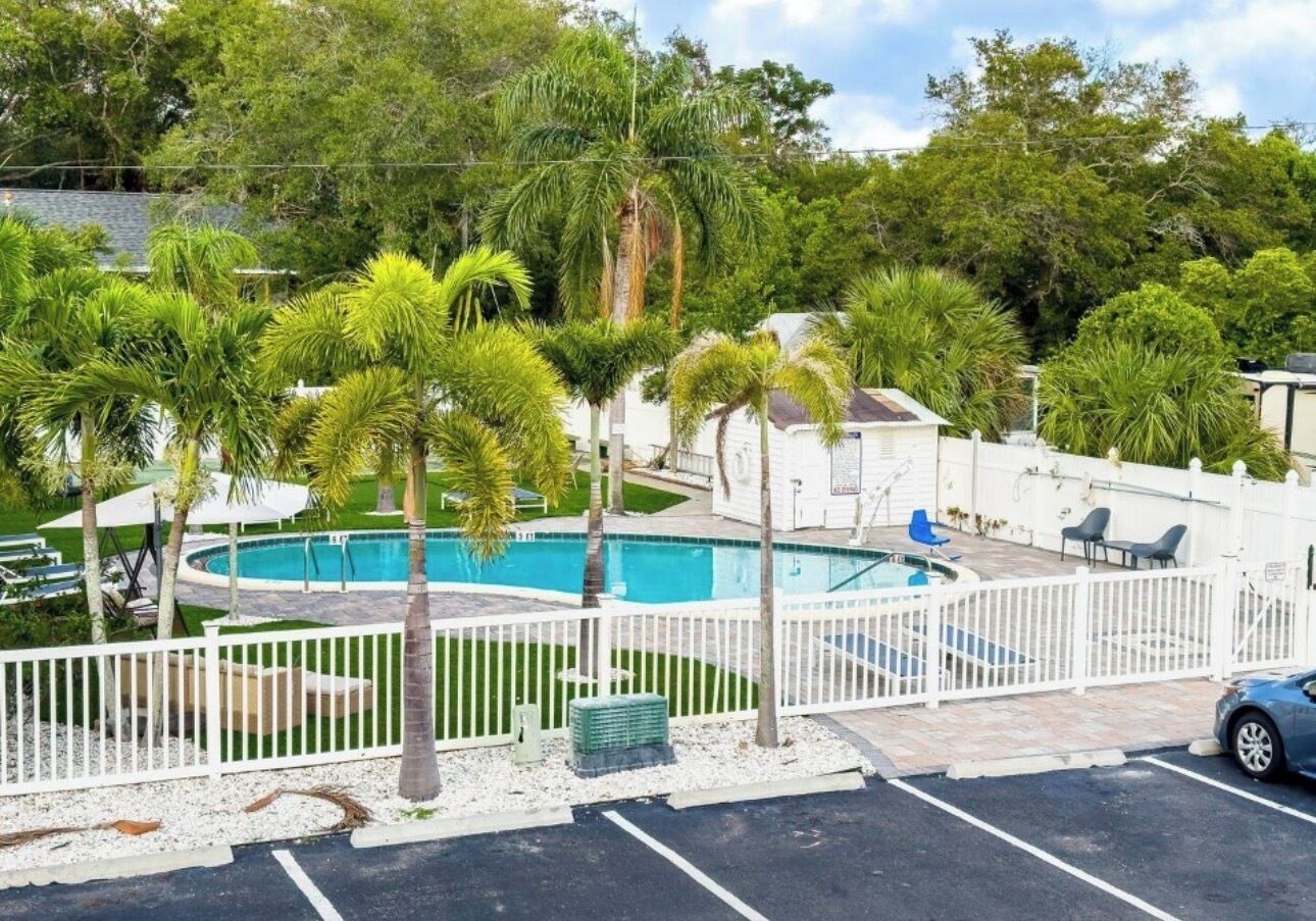 A swimming pool surrounded by palm trees and a white fence, with a car parked nearby.