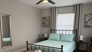 A bedroom with a bed, a ceiling fan, a window with curtains, a bedside table, a nightstand, a mirror, and a wall-mounted artwork. - Beach Retreat: Modern 1-Bedroom • Pet Friendly (Clearwater)
