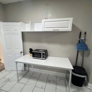 Una pequeña kitchenette con microondas, una mesa blanca y un armario blanco encima.