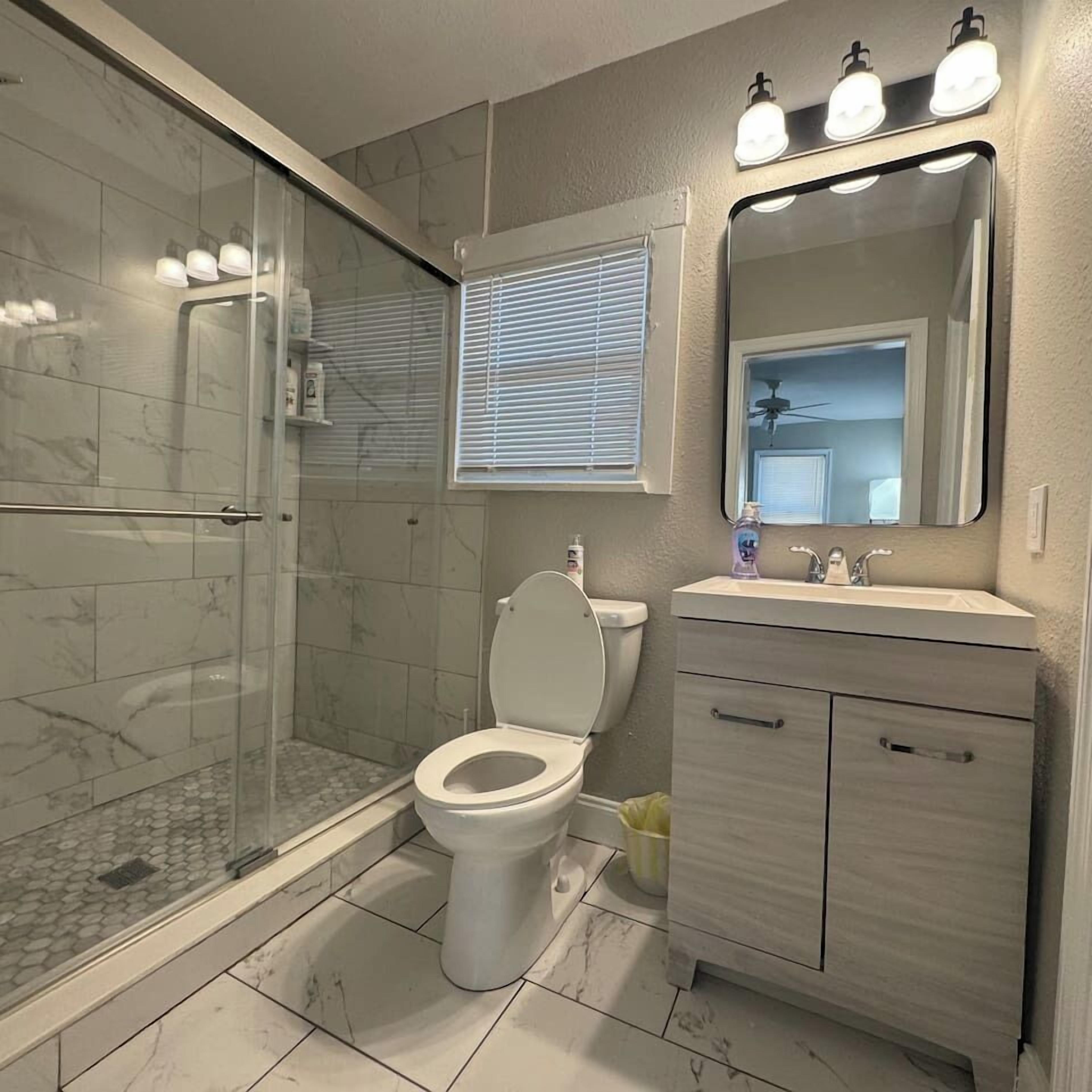 Une salle de bain avec une douche à parois de verre, des toilettes, un meuble-lavabo avec un miroir et une fenêtre avec des stores.