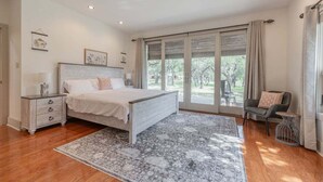 Une chambre avec un grand lit, une table de chevet, une chaise, une fenêtre avec des rideaux et une vue sur la verdure à l’extérieur.