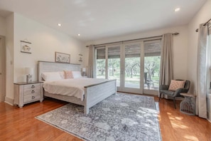 Una camera da letto con un letto grande, un comodino, una sedia, una finestra con tende e vista sul verde.