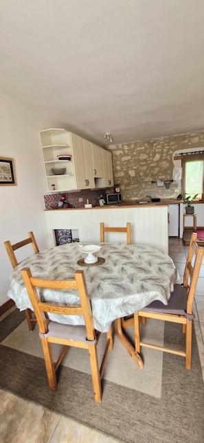 Un coin repas avec une table ronde et des chaises en bois, une cuisine avec des meubles blancs et un mur en pierre, et une fenêtre offrant une vue agréable.
