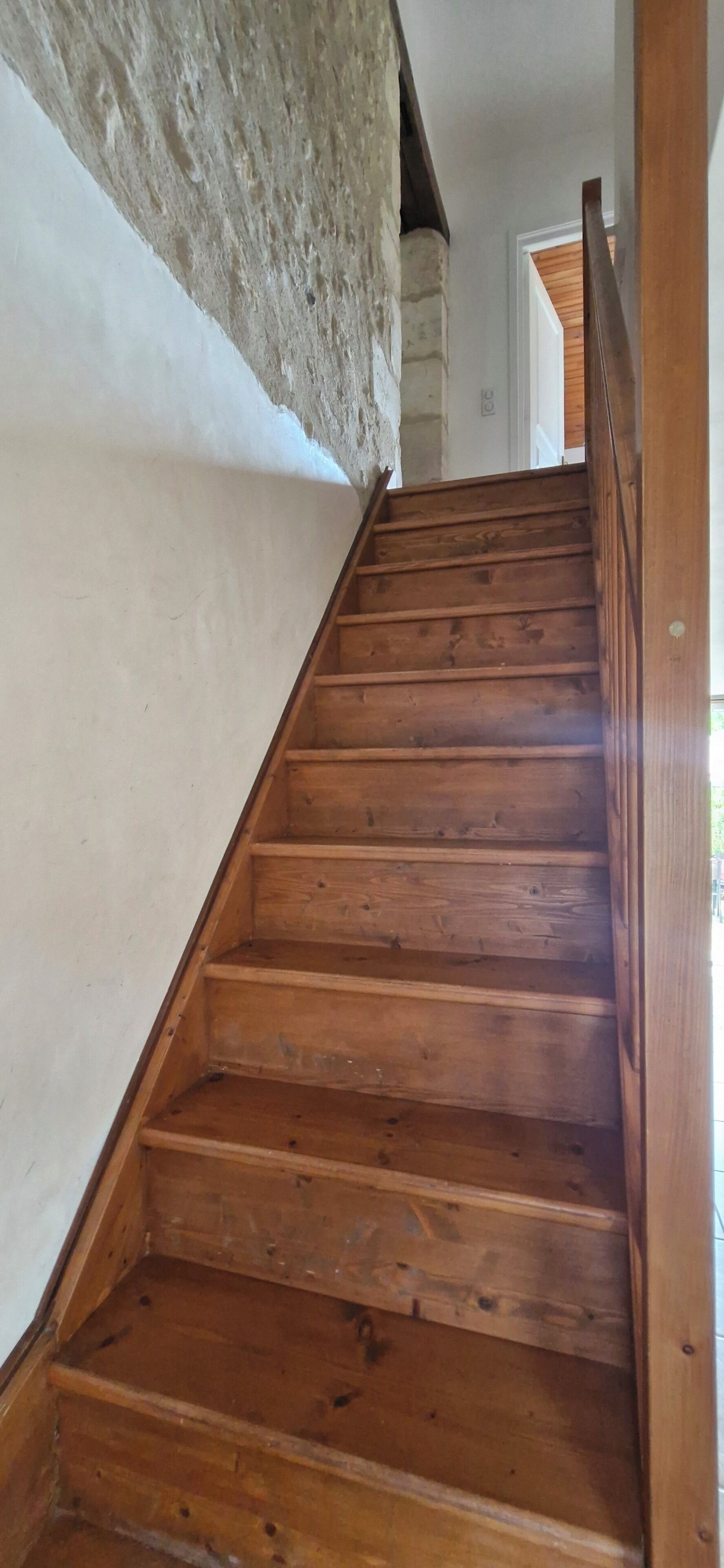 A wooden staircase with a handrail, leading to a room with a white wall and a stone wall.