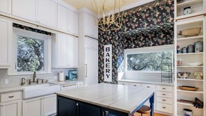 A kitchen with white cabinetry, a central island, a 'BAKERY' sign, and a window overlooking greenery. - Unique Victorian Home*Pool*Pets*Firepit (Dripping Springs)