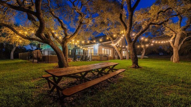 A picnic table in a grassy area with trees and string lights. - Hill Country Retreat | Private Pool | Hot Tub | Sleeps to 12 (Dripping Springs)