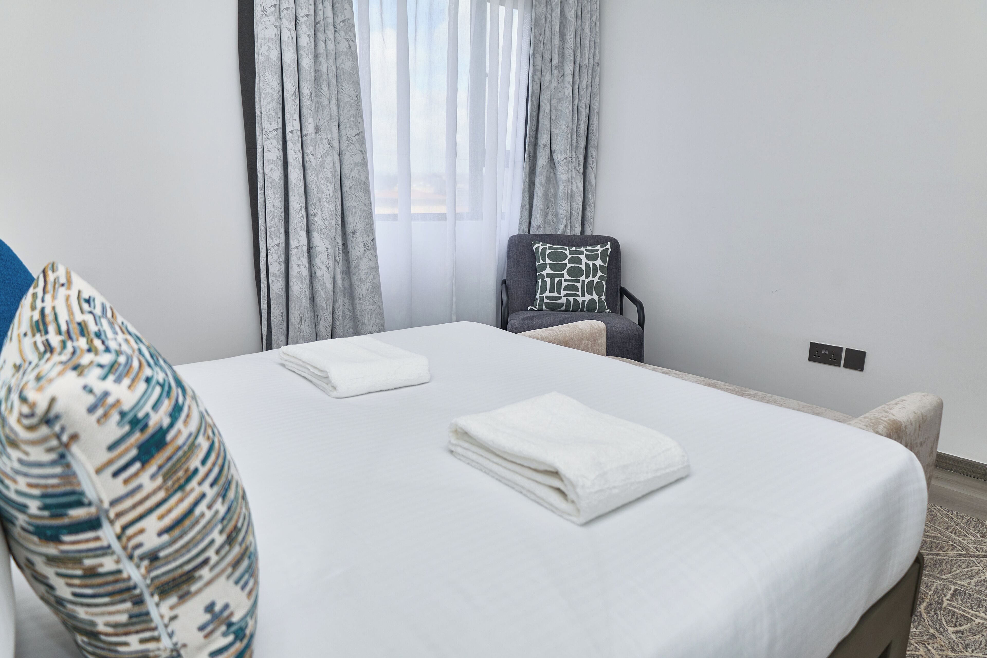 A neatly made bed with white linens, a chair with a patterned cushion, and a window with sheer curtains.