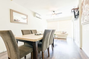 Ein Essbereich mit Holztisch und sechs Stühlen, Deckenventilator, Wandklimaanlage, einer Couch und einem Fenster mit Jalousien.