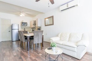 Ein modernes Wohnzimmer mit einer Couch, einem Esstisch mit Stühlen, einem kleinen runden Tisch mit Pflanze und einem Deckenventilator.