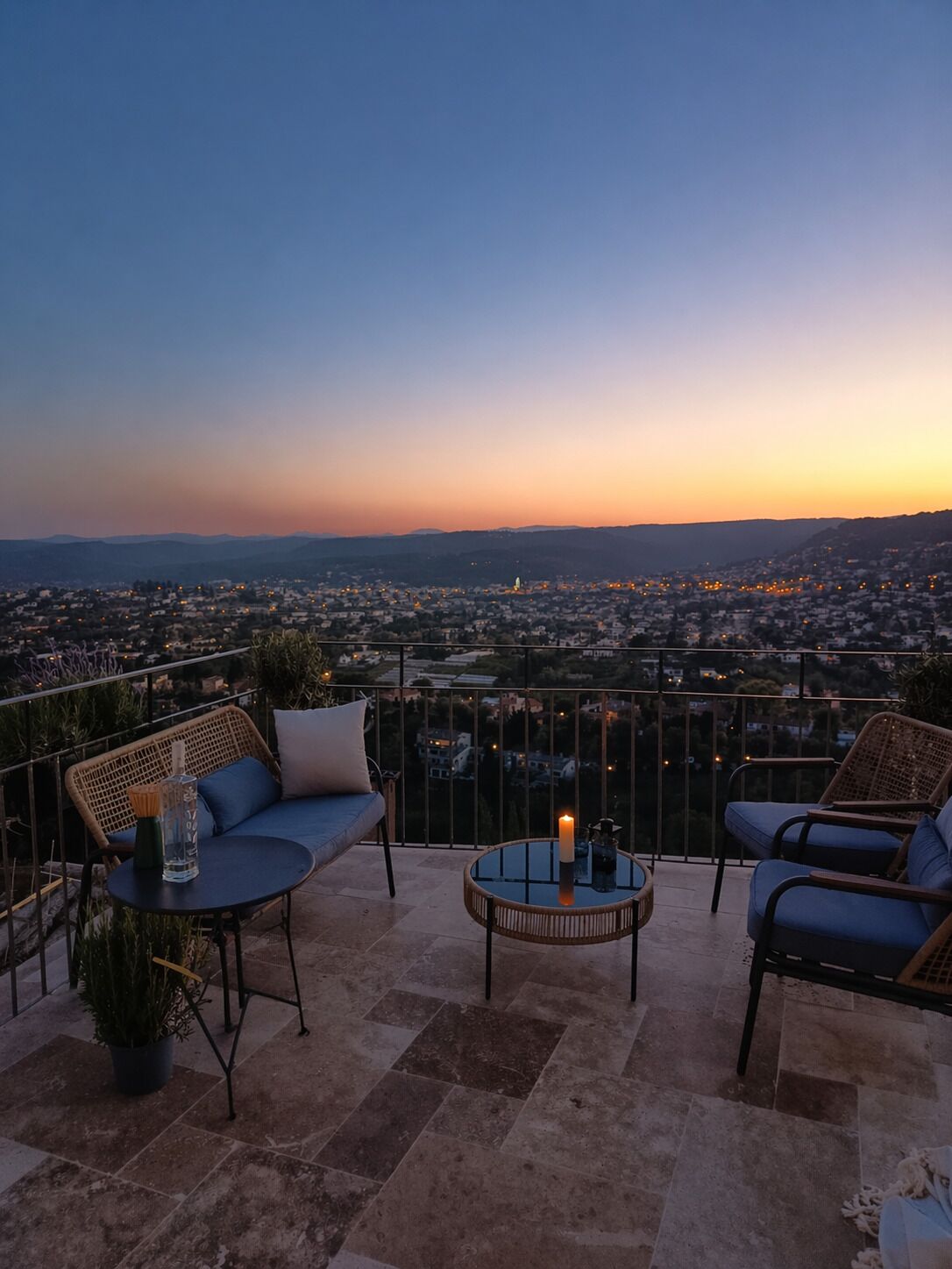 Un balcón con mobiliario de mimbre, una mesa de vidrio y vista a un paisaje urbano al anochecer.