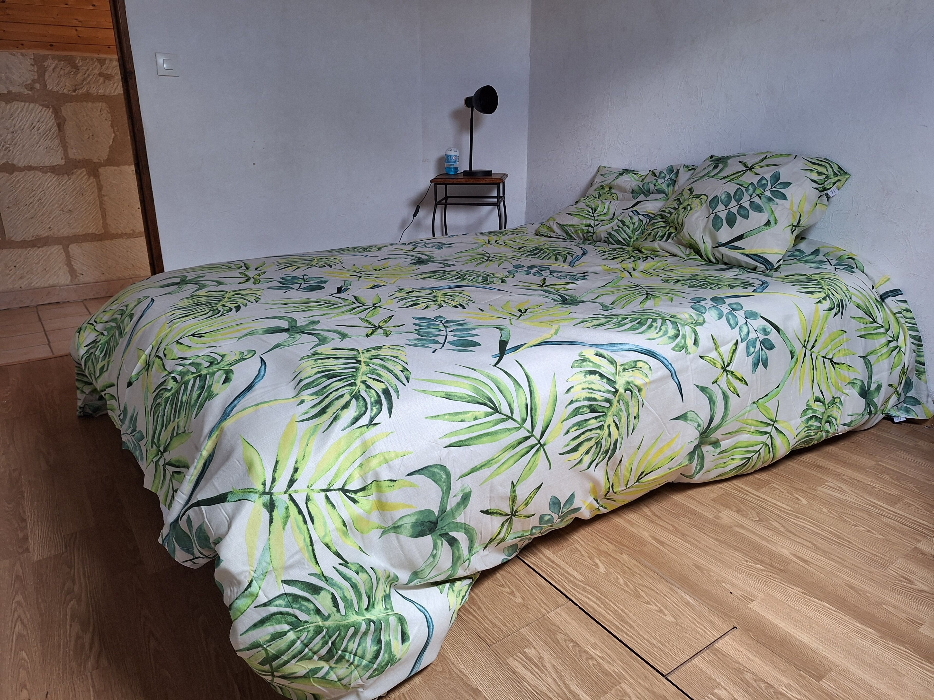 A bed with a tropical leaf pattern bedspread and matching pillows, a small bedside table with a lamp, and a stone wall.