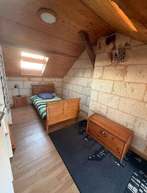A bedroom with a sloped ceiling, a wooden bed, a dresser, a small window, and a stone wall.