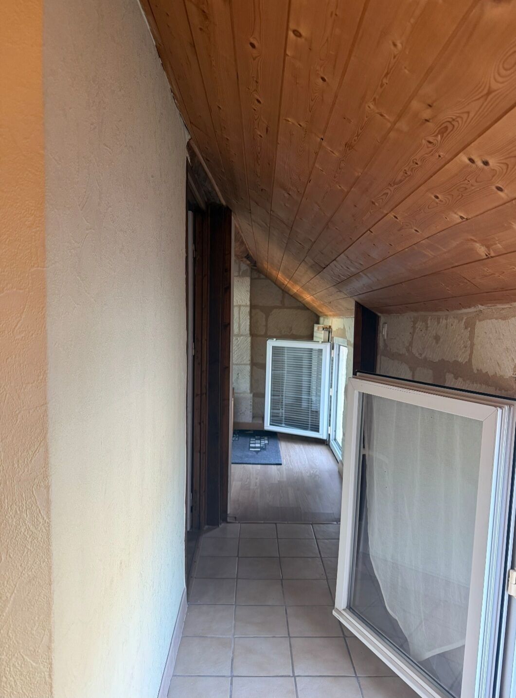 Een smalle gang met houten plafond en betegelde vloer, die leidt naar een kamer met een groot raam en een deur.