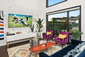 Ein modernes Wohnzimmer mit großem Fernseher, violetten Sesseln, einem blauen Sofa und einem Holztisch.