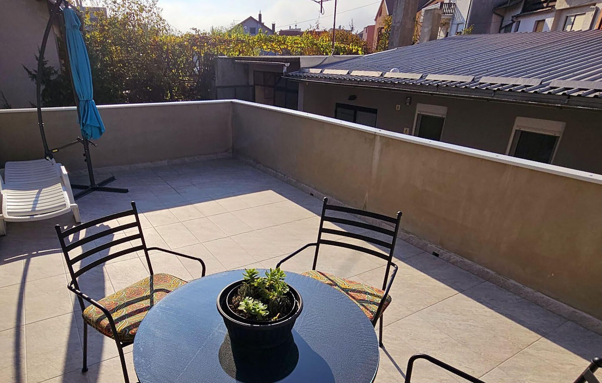 Une terrasse sur le toit avec une table ronde, deux chaises, un pot de fleurs et un parasol bleu.