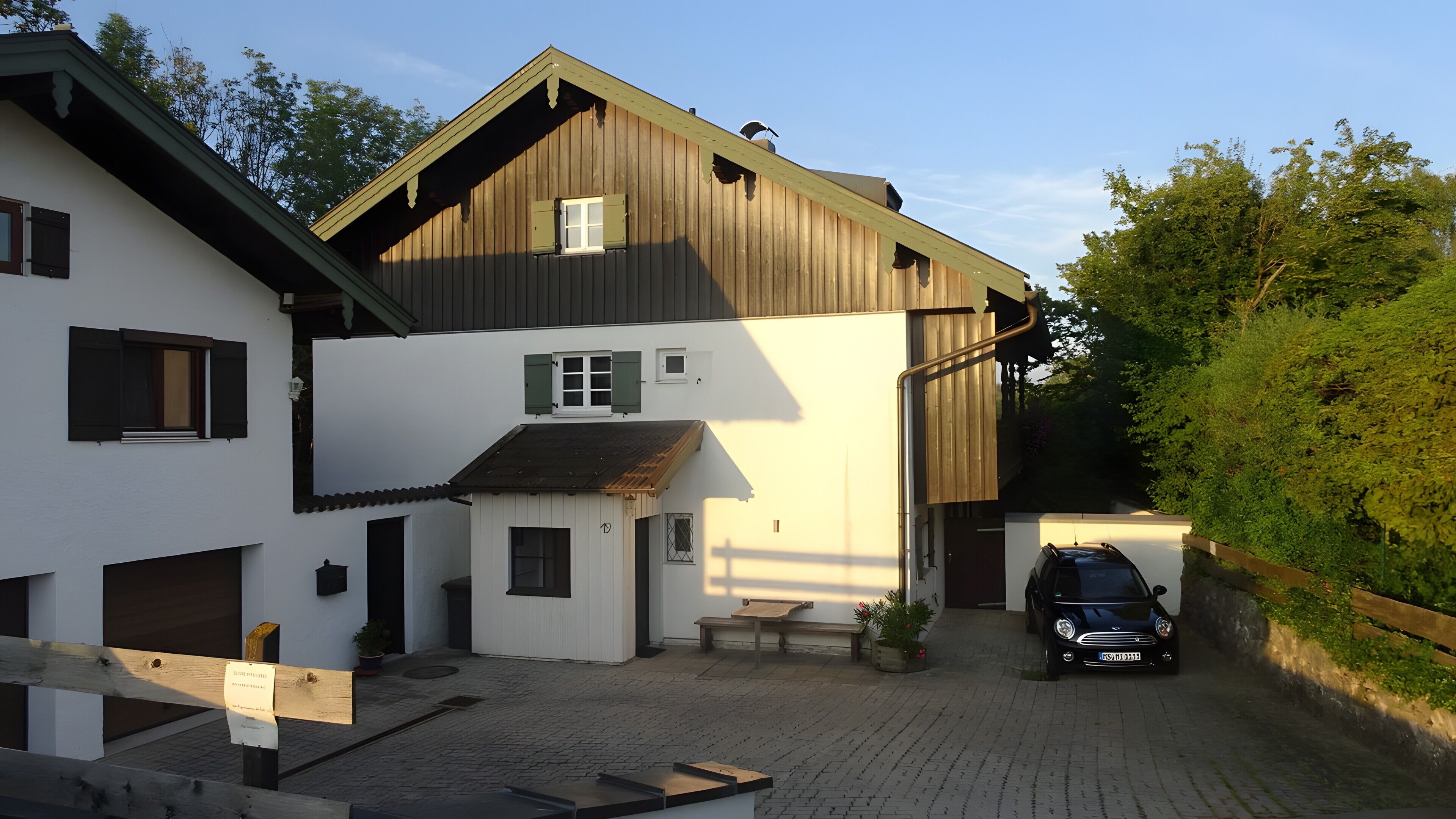 A two-story building with a sloped roof, a car parked in the driveway, and a paved courtyard.