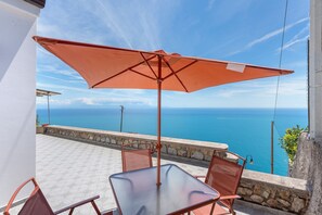 A terrace with a table and chairs, a large umbrella, and a view of the sea. - Olimpo Holiday Home with Sea and Mountain Views (Conca dei Marini)