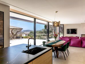 A modern kitchen with a large window, a dining area with a table and chairs, and a living room with a sofa. - Lux CostaBravaSi - Luxury, design, sustainability (Platja d'Aro)
