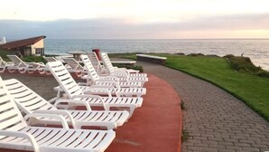 Fileira de cadeiras de praia brancas ao longo de um caminho de paralelepípedos, próximo ao mar, com uma área gramada e um edifício ao fundo.