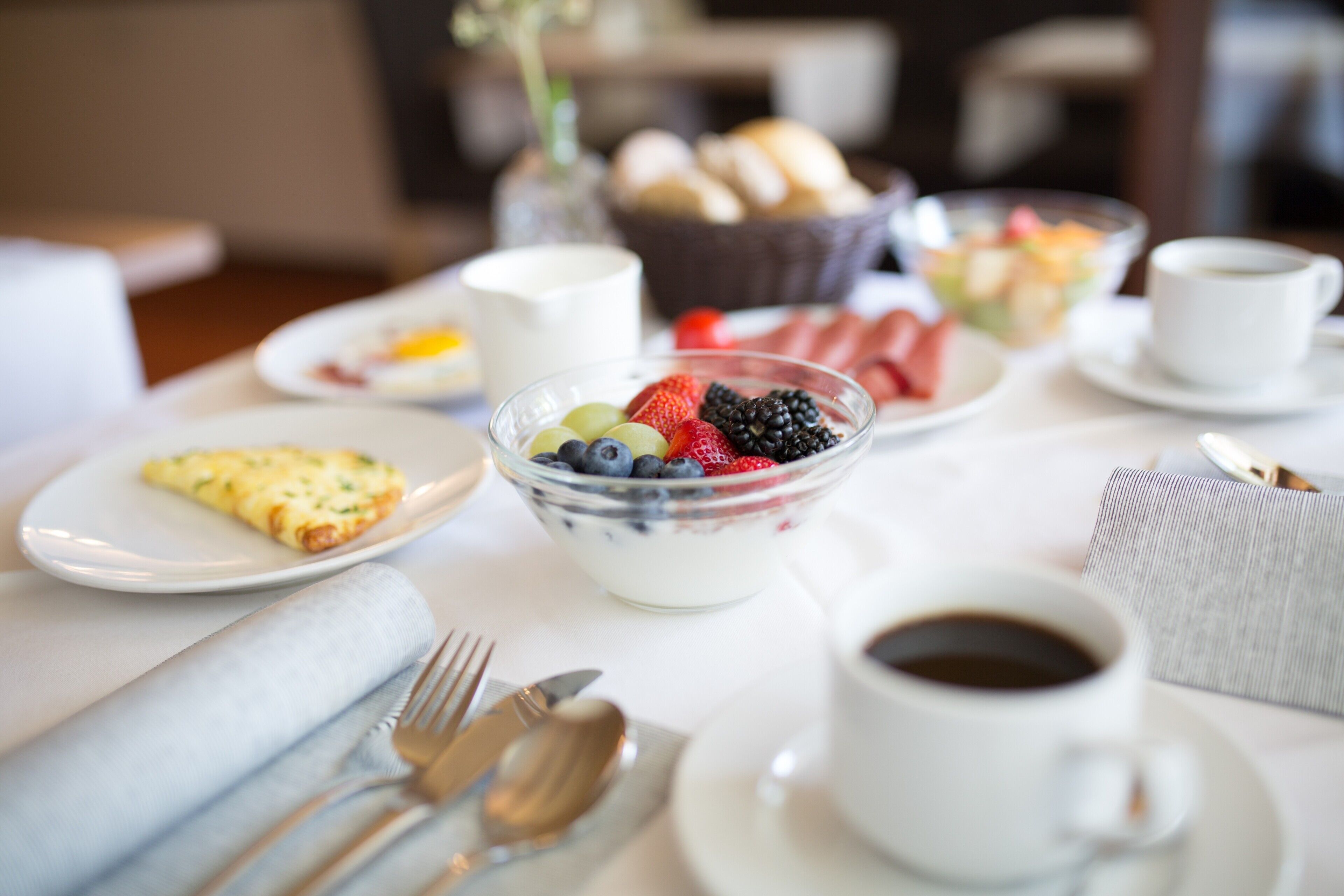 A breakfast setting with a bowl of yogurt and berries, a plate of toast, a cup of coffee, and a basket of bread.