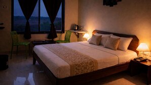 A hotel room with a bed, two chairs, a nightstand with a lamp, and a view of the outside. - Varad International Hotel and Banquet (Alapur)