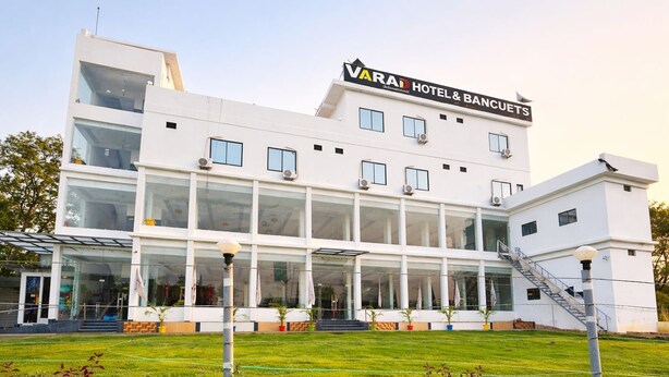 A modern hotel with a glass facade, a prominent sign reading 'VARAD HOTEL & BANCUETS,' and a well-maintained lawn in front. - Varad International Hotel and Banquet (Alapur)