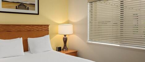 A hotel room with a bed, a nightstand with a lamp, a picture on the wall, and a window with blinds.