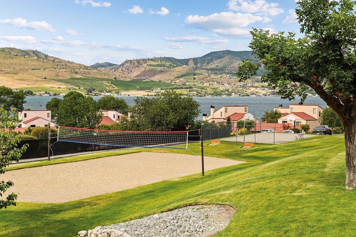 Cancha de voleibol con red, rodeada de césped, con casas y un lago al fondo.