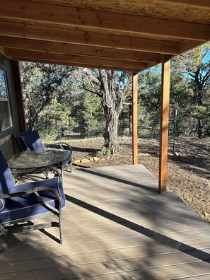 A covered outdoor seating area with a table and chairs, wooden beams, and a view of trees and a fence.
