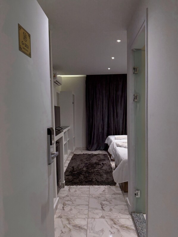 A narrow hallway leading to a bedroom with a bed, a dark curtain, a small desk, and a mounted air conditioner. - DHB STAYS (Cairo)